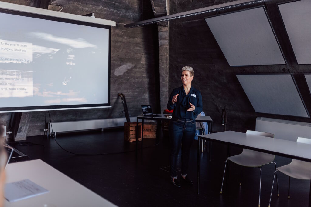 Marta Weiss presents at the Future is WOW, wearing a dark shirt and jeans, with a powerpoint presentation behind her.