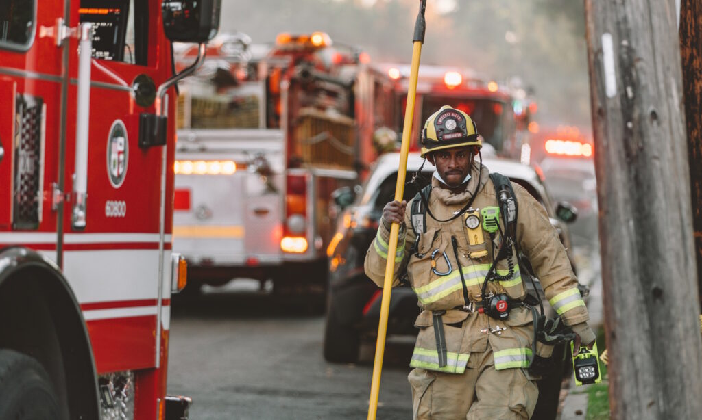 Firefighter in the foreground with fire trucks and smoke in the background.