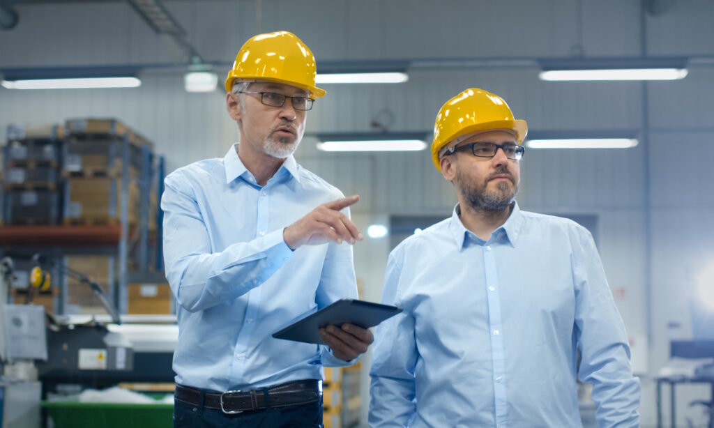 Two men in pale blue shirts and yellow helmets point to potential hazards in a factory.