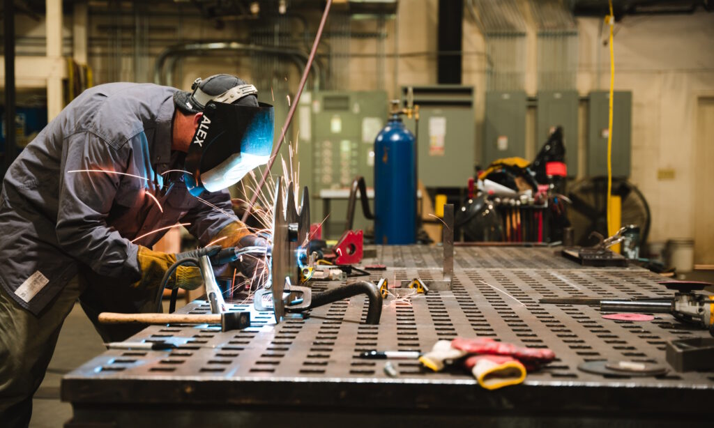 A worker wearing PPE and face shield welding at a workbench, sparks flying.