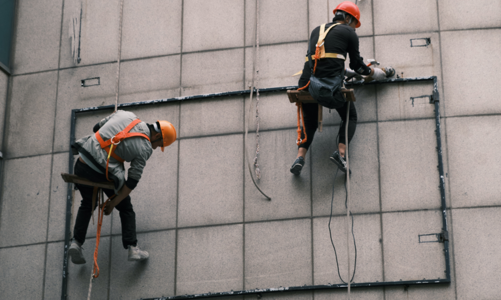 Two people suspended in climbing harnesses and wearing helmets work on a concrete wall.