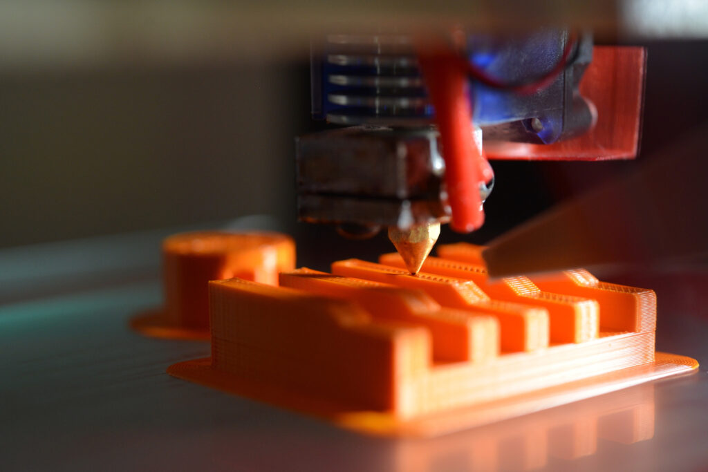 3D printer making an orange plastic component.