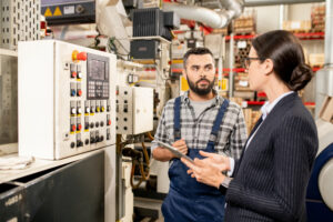 Maintenance worker in grey checkered shirt and blue dungarees discusses a piece of equipment full of buttons with a woman in a black blazer and white shirt, holding a tablet.