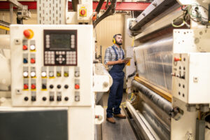 A maintenance worker, wearing a grey checkered shirt, blue dungarees and yellow ear defenders holds a tablet and watches a piece of machinery creating sheets of see-through plastic film.