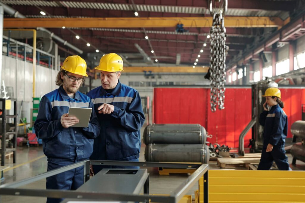 Two men wearing blue overalls and yellow hard hats review a permit to work on a mobile device whilst standing in front of machinery.