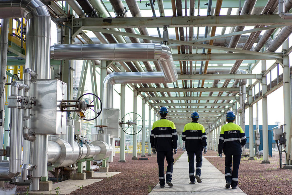 Three engineers in high-vis and blue overalls and blue hardhats walk beneath metal piping at a chemical site.