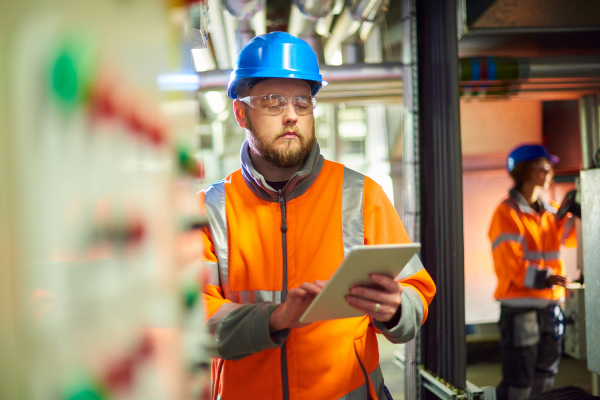 Male industrial worker wearing PPE and using a tablet to perform real-time inspections or safety checks on the factory floor.